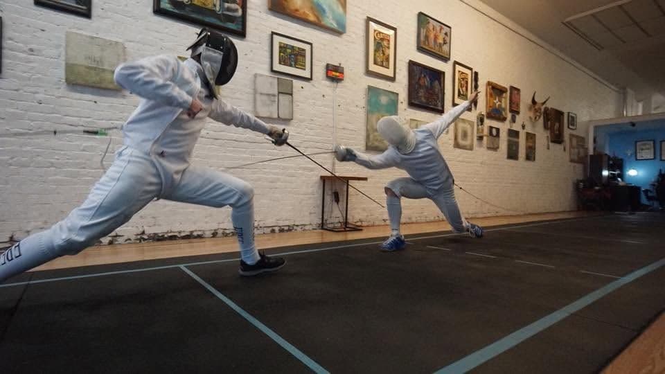 Two masked fencers at full extension on the piste at Marin Fencing Academy, beneath the salle's wall of prints