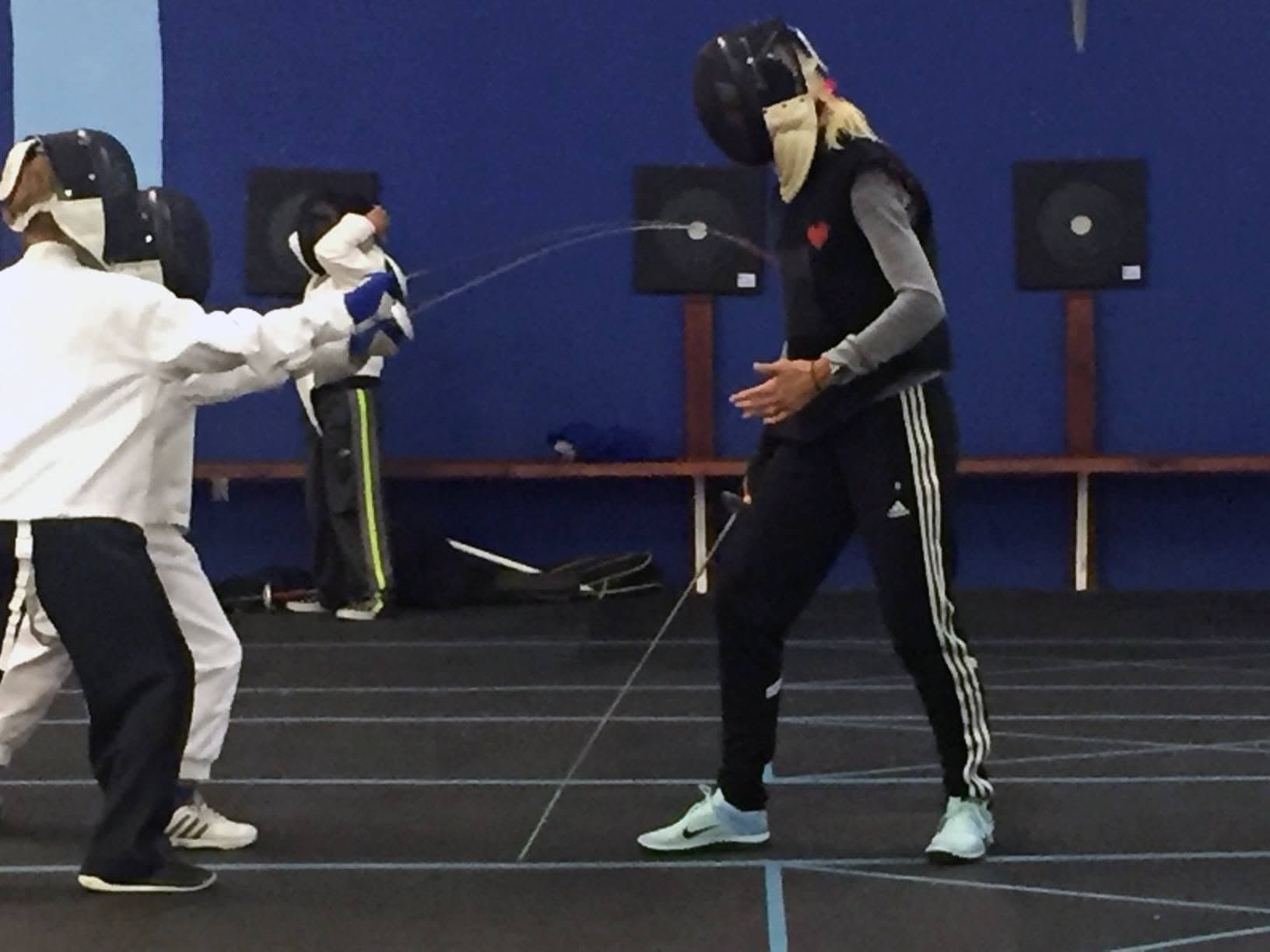 A coach in whites teaching a masked beginner in ordinary clothes, the blade bending on a hit