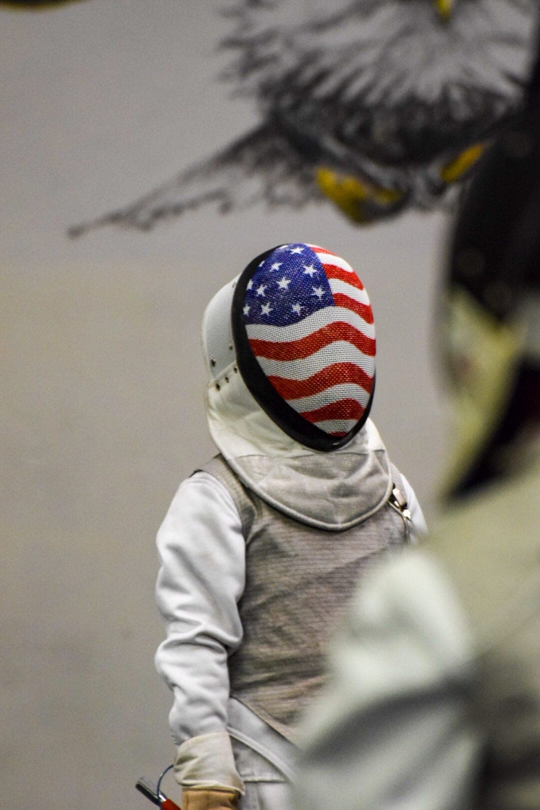 A masked fencer standing ready, beneath the club’s painted eagle